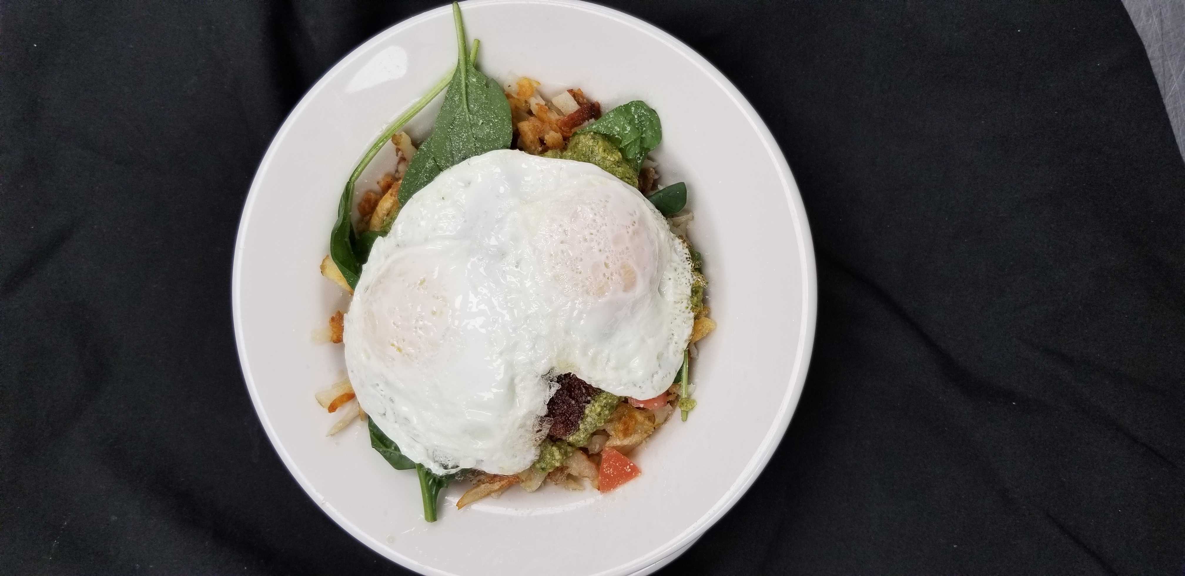 Analog film overhead of fried eggs over hash and vegetables on a white plate, golden yolk and olive-green tones, dark cloth backdrop.