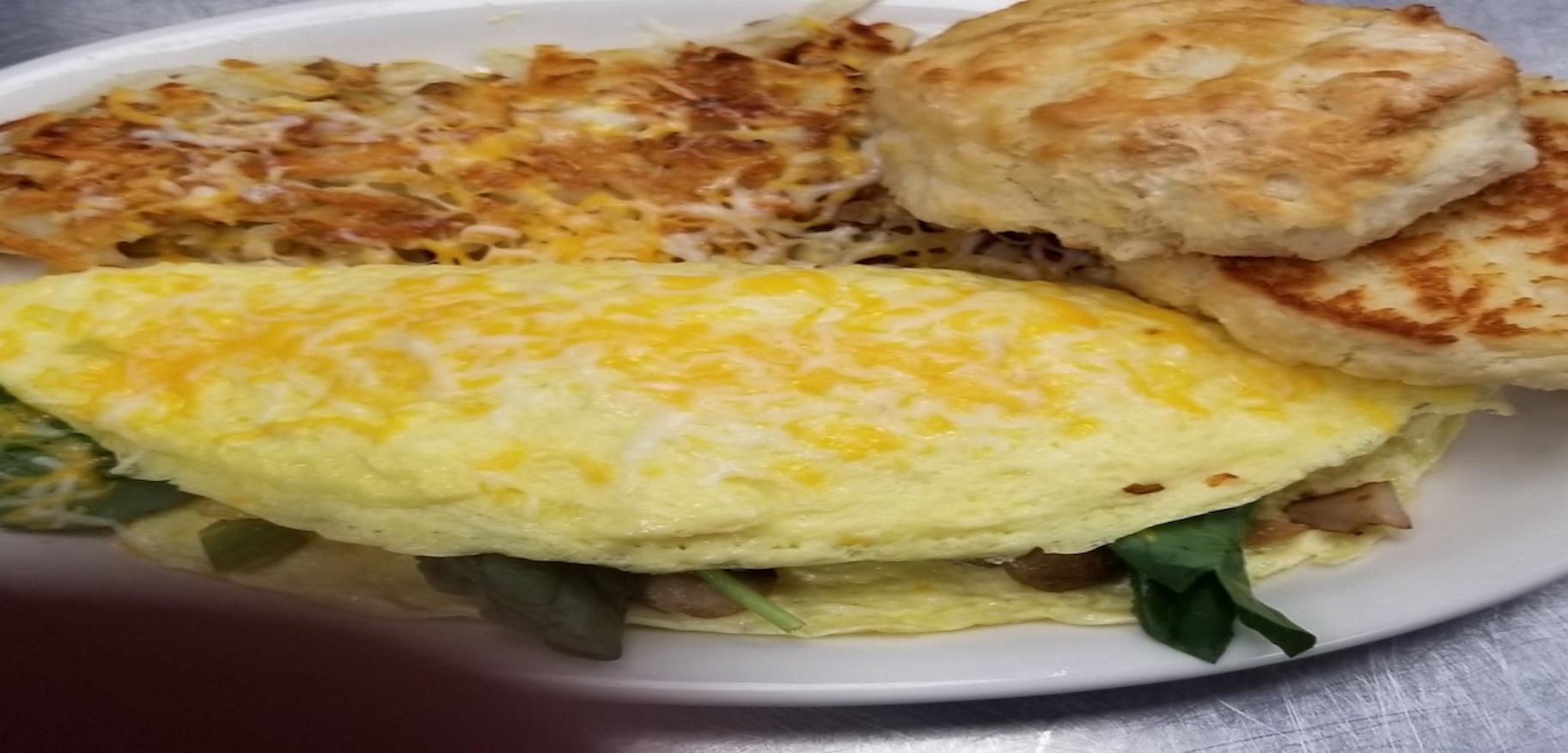 Analog film overhead of a cheese-topped omelet with spinach beside hash browns and biscuits, golden-yellow and green tones.